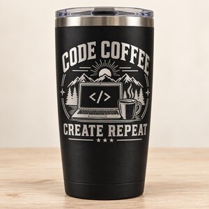 May include: A matte black stainless steel tumbler with a clear lid. The front features a white laser-engraved graphic of a laptop with code symbols on the screen, a coffee mug, mountains, and trees, with the text CODE COFFEE CREATE REPEAT.