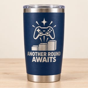 May include: A dark blue stainless steel tumbler with a clear lid. The front features a laser-engraved graphic of a video game controller above a three-tiered podium, with the text "ANOTHER ROUND AWAITS" printed in a bold, sans-serif font.