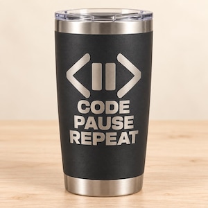 May include: A matte black stainless steel tumbler with a clear lid and a silver metallic rim at the base. The front features an engraved design with two angle brackets surrounding a pause symbol, followed by the text CODE PAUSE REPEAT.