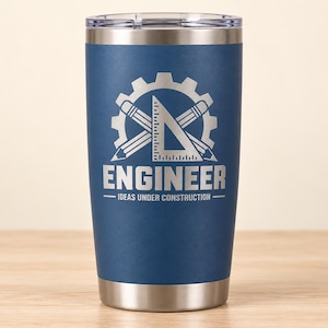 May include: A navy blue stainless steel tumbler with a clear lid. The front features a laser-engraved design of a gear, two pencils, and a triangle ruler, with the text "ENGINEER" and "IDEAS UNDER CONSTRUCTION" printed in a bold font.