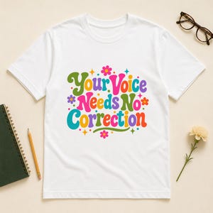 May include: A white short-sleeve t-shirt featuring the colorful text "Your Voice Needs No Correction" in a retro font, surrounded by small flower and star graphics in shades of pink, orange, yellow, green, blue, and purple.