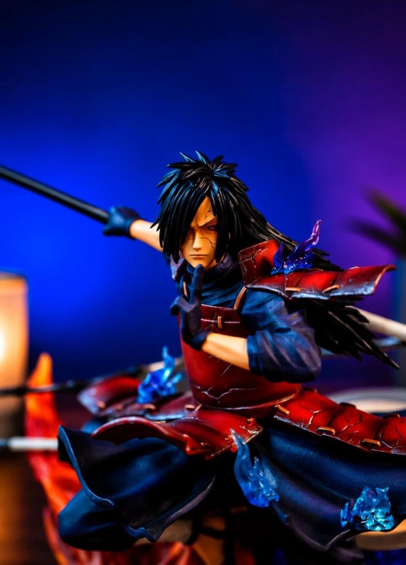 May include: A detailed anime character figurine featuring a figure with long black hair, wearing dark blue clothing and red segmented armor. The character is posed in a dynamic stance with one hand raised, accented by translucent blue flame effects.