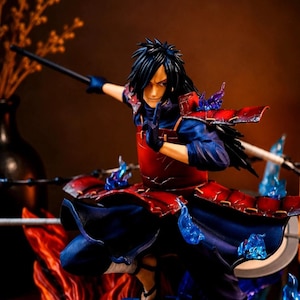 May include: A detailed anime character figurine featuring a figure with long black hair, wearing red segmented armor over a dark blue outfit. The figure is posed in a dynamic stance with one hand raised, surrounded by translucent blue flame effects.