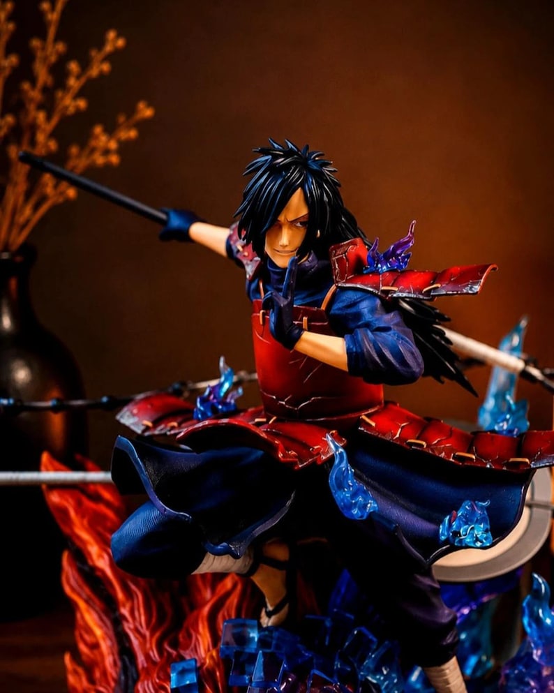 May include: A detailed anime character figurine featuring a figure with long black hair, wearing red segmented armor over a dark blue outfit. The figure is posed in a dynamic stance with one hand raised, surrounded by translucent blue flame effects.