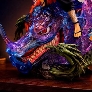 May include: A detailed anime-style figurine featuring a translucent blue and purple dragon head with a sharp horn and visible teeth. A human foot wearing a black sandal rests on the dragon, which is accented by green textured elements.