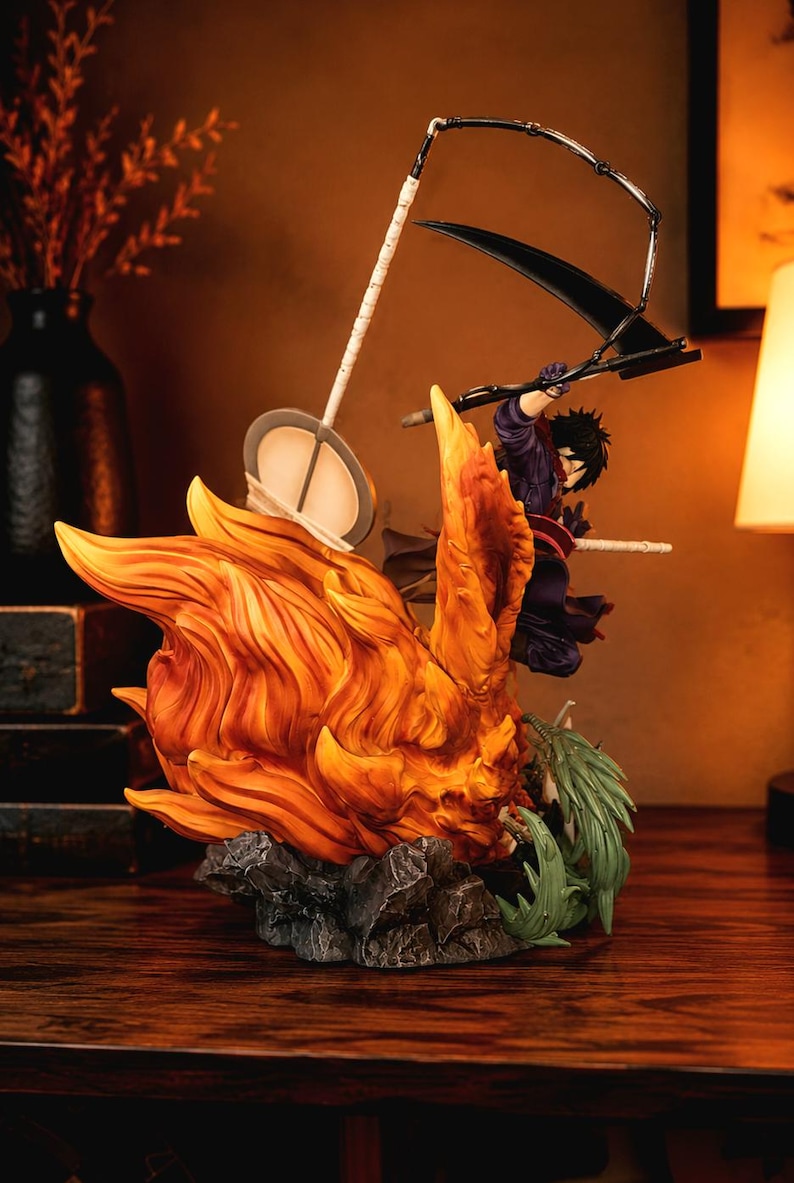 May include: A dynamic anime-style figurine featuring a character with dark hair in a purple outfit, wielding a large black scythe. The figure is surrounded by sculpted orange flames and green foliage, set upon a textured gray rocky base.