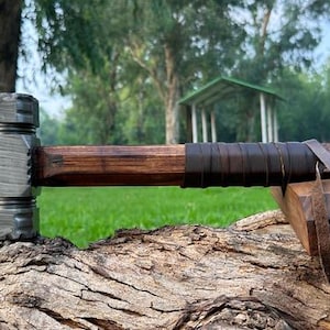 Könnte beinhalten: Ein Hammer mit einem gemusterten Damaststahlkopf, der eine geschichtete metallische Textur aufweist. Der Griff ist aus dunkelbraunem Holz gefertigt, mit einem dunklen Lederband am Ende umwickelt und hat eine dünne Lederkordel am Griff.