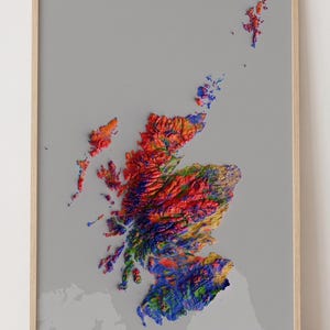 May include: A framed topographic map of Scotland featuring a vibrant, multi-coloured relief design. The landmass is rendered in shades of red, orange, green, blue, and yellow against a solid grey background, all enclosed in a light wood frame.