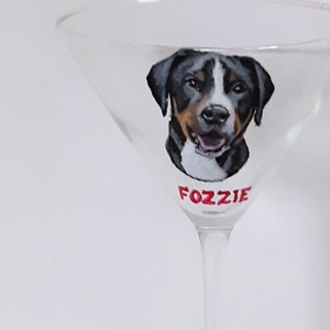 May include: A clear glass martini glass with a painted image of a black, white, and brown dog with the name "FOZZIE" written in red underneath the image.