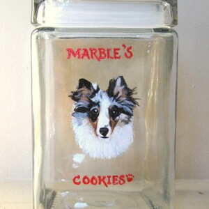 Op de afbeelding: Een glazen koekjespot met een geschilderd beeld van een hond en de tekst "Marble's Cookies".