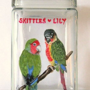 May include: A clear glass jar with a painted design of two green and red parrots on a branch. The jar has the words "Skittles ♥ Lily" painted on the front.