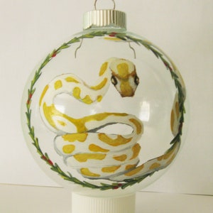 Op de afbeelding: Een transparante glazen kerstbal met een geschilderd beeld van een gele en witte slang erin. De kerstbal is versierd met een groene hulstkrans en rode bessen.