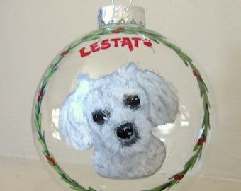 Puede incluir: Adorno navideño de cristal transparente con un retrato de perro blanco pintado a mano. El perro tiene ojos y nariz negros. El nombre "LESTAT" está escrito en rojo encima del perro. Una corona verde con bayas rojas rodea al perro.