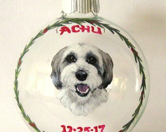 Puede incluir: Un adorno de Navidad de vidrio transparente con un retrato pintado a mano de un perro blanco y gris. El adorno está decorado con una corona verde y bayas rojas. El nombre del perro, "ACHU", y la fecha "12-25-17" están pintados en el adorno.