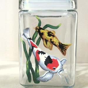 May include: Hand-painted glass jar with a lid featuring two koi fish swimming among green aquatic plants. The fish are painted in shades of red, white, black, and yellow.