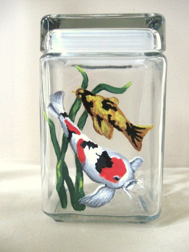 May include: Hand-painted glass jar with a lid featuring two koi fish swimming among green aquatic plants. The fish are painted in shades of red, white, black, and yellow.
