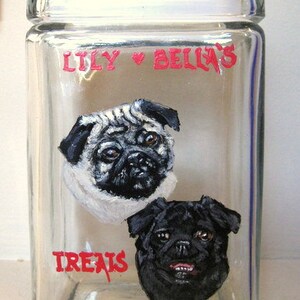 Puede incluir: Un tarro de vidrio transparente con una inscripción pintada en rojo "Lily & Bella's Treats". El tarro presenta dos perros pug pintados, uno blanco y uno negro.