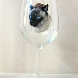 Può includere: Un bicchiere di vino in vetro trasparente con un'immagine dipinta di un gatto siamese con un cappello marrone. Il gatto ha gli occhi azzurri e un manto bianco e marrone chiaro.