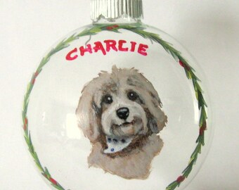 Puede incluir: Un adorno de Navidad de vidrio transparente con un retrato pintado a mano de un perro. El perro es marrón claro con un pecho blanco y una bandana azul. El adorno está decorado con una corona de acebo verde y roja y el nombre "Charlie" está escrito en rojo.