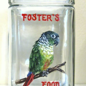 May include: A clear glass jar with a red painted label that says "FOSTER'S FOOD". A green and red parakeet is painted on the jar, perched on a branch.
