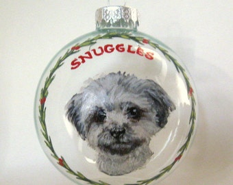 Puede incluir: Adorno navideño de cristal transparente con un retrato de perro pintado a mano. El perro tiene pelo gris y blanco. La palabra "SNUGGLES" está escrita en rojo encima del perro, rodeada por una corona verde.