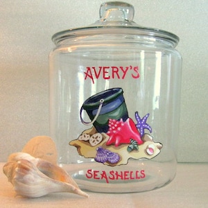 May include: A clear glass jar with a lid, decorated with a seashell theme. The jar has the words "Avery's Seashells" painted on it. The design includes a bucket, seashells, and a starfish.