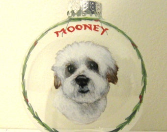 Puede incluir: Un adorno de Navidad de vidrio transparente con un perro blanco y marrón y el nombre "Mooney" escrito en la bola. La bola está adornada con una corona de acebo verde.