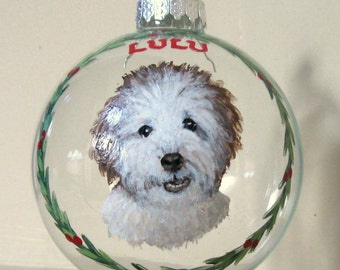 Puede incluir: Un adorno de Navidad de vidrio transparente con una imagen pintada de un perro blanco con pelo marrón. El adorno tiene una parte superior plateada y una corona de acebo verde con bayas rojas alrededor del borde. El nombre del perro, "LULU", está pintado en rojo en el adorno.