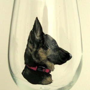 Puede incluir: Una copa de vino transparente con una imagen pintada a mano de un perro pastor alemán. El perro se representa de perfil, con pelaje negro, marrón y canela, y un collar rojo. La copa está sobre un fondo blanco.