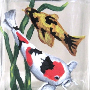 May include: Hand-painted glass block featuring two koi fish swimming among green aquatic plants. The larger fish is white with black and red markings, while the smaller fish is yellow with black markings.