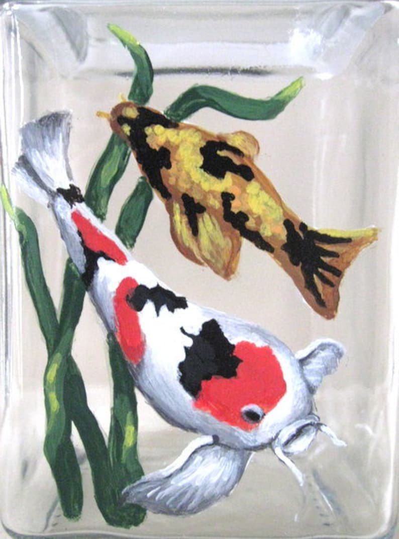 May include: Hand-painted glass block featuring two koi fish swimming among green aquatic plants. The larger fish is white with black and red markings, while the smaller fish is yellow with black markings.