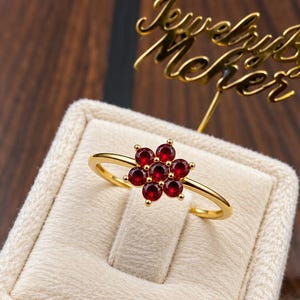 May include: A gold ring featuring a flower-shaped cluster of seven round, deep red gemstones. The ring is displayed on a beige velvet ring box, with a gold-colored sign behind it that reads JewelryByMeher in cursive script.
