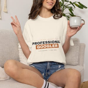May include: An off-white crewneck t-shirt featuring the text "JOB TITLE: UNOFFICIAL PROFESSIONAL GOOGLER" in black and brown, with the additional text "0.42 seconds, 4 tabs deep" printed below in a smaller black font on the front of the shirt.
