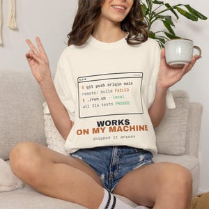 May include: An off-white short-sleeve t-shirt featuring a graphic of a terminal window with the text "git push origin main", "build FAILED", "./run.sh --local", and "all 214 tests PASSED". Below is the text "WORKS ON MY MACHINE" and "shipped it anyway".