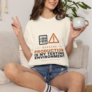 May include: An off-white short-sleeve t-shirt featuring a graphic with a server icon, a warning triangle, and the text: WARNING PRODUCTION IS MY TESTING ENVIRONMENT staging is a state of mind. The shirt is paired with blue denim shorts.