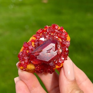 Op de afbeelding: Een levendig rood kristallijn mineraal exemplaar met een grote, centrale, gefacetteerd uitziende kristal omringd door kleinere, gegroepeerde rode kristallen op een getextureerde, geelbruine matrixbasis met diepe karmozijnrode tinten.