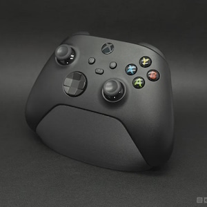 May include: A matte black wireless video game controller featuring two analogue sticks, a directional pad, and four action buttons labelled with the letters A, B, X, and Y in green, red, blue, and yellow colours respectively, centred on a dark surface.