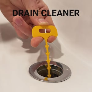 May include: A bright yellow plastic drain cleaner tool with a flat handle and a long, thin stem featuring multiple small, upward-pointing barbs along its length. The text DRAIN CLEANER is printed in black above the tool, which is inserted into a drain.