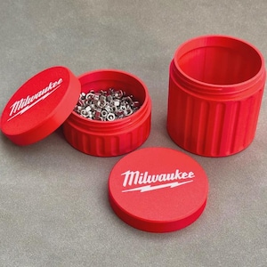 May include: Two red cylindrical storage containers with ribbed sides and matching lids. Each lid features the white Milwaukee logo. One container is open and filled with small silver metal nuts, while the other container stands empty next to it.