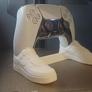 May include: A white video game controller stand featuring two miniature white trainers as the base. The controller is white with black accents, including two analogue sticks and a central touch pad. The trainers are detailed with laces and a side swoosh.
