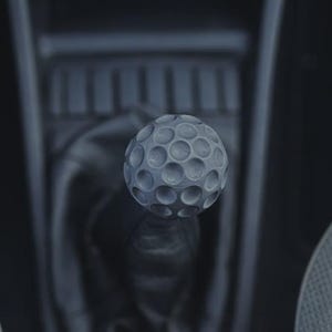 May include: A spherical gear shift knob with a textured dimpled surface resembling a golf ball. The knob is matte grey and mounted on a black leather-like boot within a vehicle interior, positioned above a dark plastic centre console area.