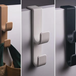 May include: Three over-the-door hooks in tan, white, and black. Each hook features a vertical ribbed texture, a flat top bracket for mounting over a door edge, and two upward-curving hooks for hanging items. A green bag hangs from the tan hook.