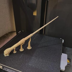 May include: A light beige 3D printed wand featuring a textured, wood-like handle with a gnarled, root-inspired base. The wand is long, slender, and tapers to a point, supported by three small, thin vertical stands attached to the main body.