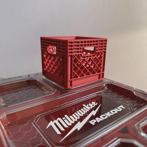 May include: A small red plastic crate with a lattice design and integrated handles, positioned on top of a clear Milwaukee Packout storage container lid featuring a hexagonal pattern and the white Milwaukee Packout logo printed on the surface.