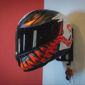 May include: A full-face motorcycle helmet featuring a custom graphic design with black, white, and vibrant red claw-like patterns. The helmet has a dark tinted visor and is mounted on a black wall bracket with a small tag reading READY TO RACE.