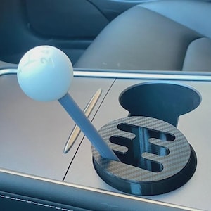 May include: A white spherical gear shift knob with a printed shift pattern sits atop a grey stick, which is inserted into a black carbon fibre patterned circular gate plate designed to fit into a vehicle cup holder console area.
