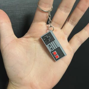 May include: A rectangular keyring shaped like a retro video game controller. The controller is grey with a black directional pad, two red circular buttons, and two small black rectangular buttons. It is attached to a silver metal split ring.