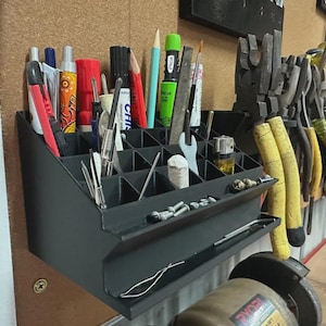 May include: A black wall-mounted organiser with multiple square compartments holding various tools, including pens, markers, a small wrench, and a lighter. The front features a shallow tray containing small metal screws and hardware components.