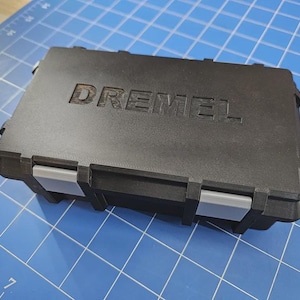 May include: A rectangular black storage case with the word DREMEL embossed on the lid. The case features a textured matte finish and two light grey plastic latches on the front edge, designed for securing the lid to the base of the container.