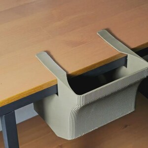 May include: A sage green desk organiser with a ribbed texture and two flat arms that hook over the edge of a tabletop. The container has a rectangular shape with curved sides and is constructed from a solid, matte-finished material.